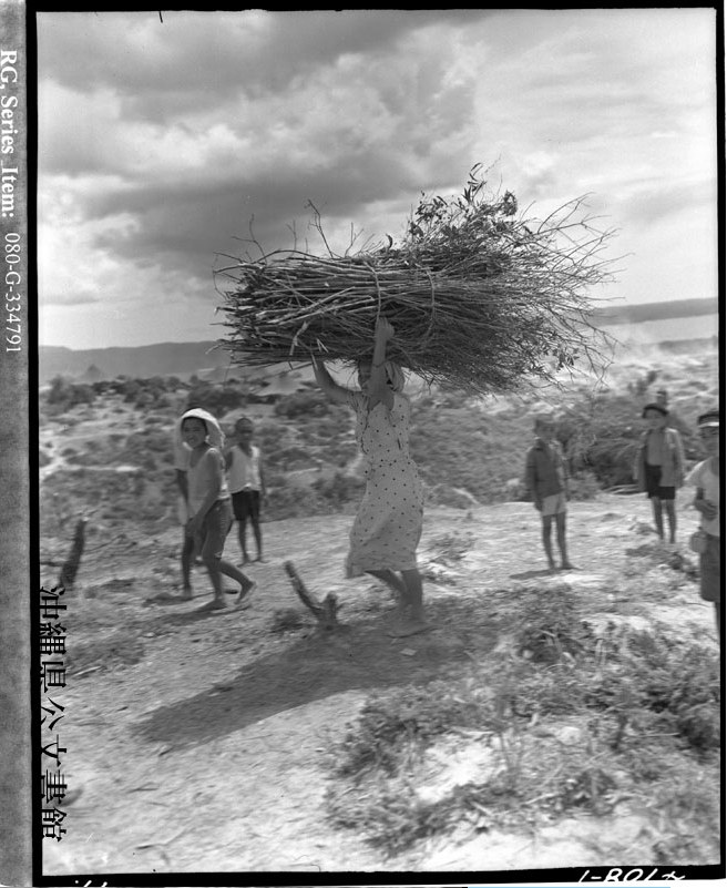 Mulher okinawana carregando gravetos na cabeça, com crianças ao redor, no campo de internamento da Baía de Oura, em Henoko, 1945.